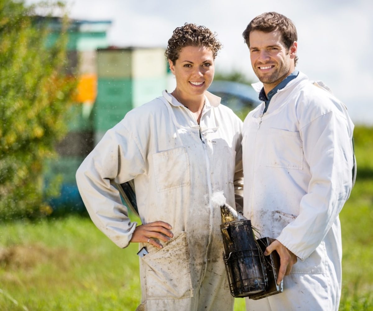 Portrait of happy male and female beekeepers with smoker standing at apiary